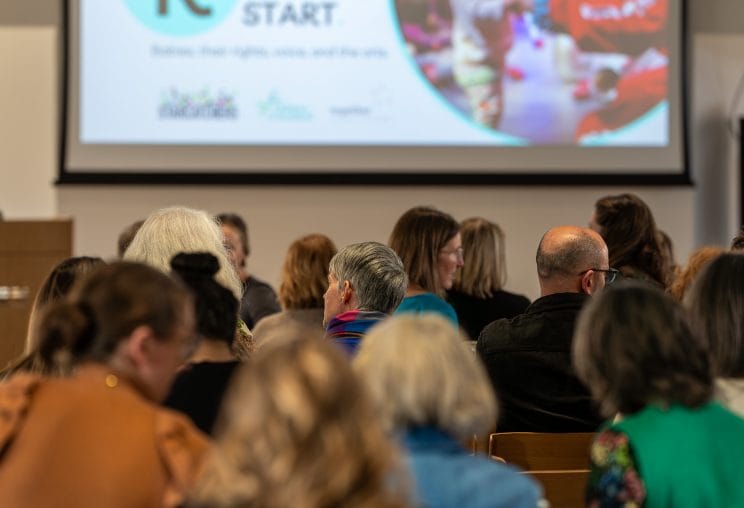 A room of conference delegates looking at a big screen with text and image showing a baby and artist together