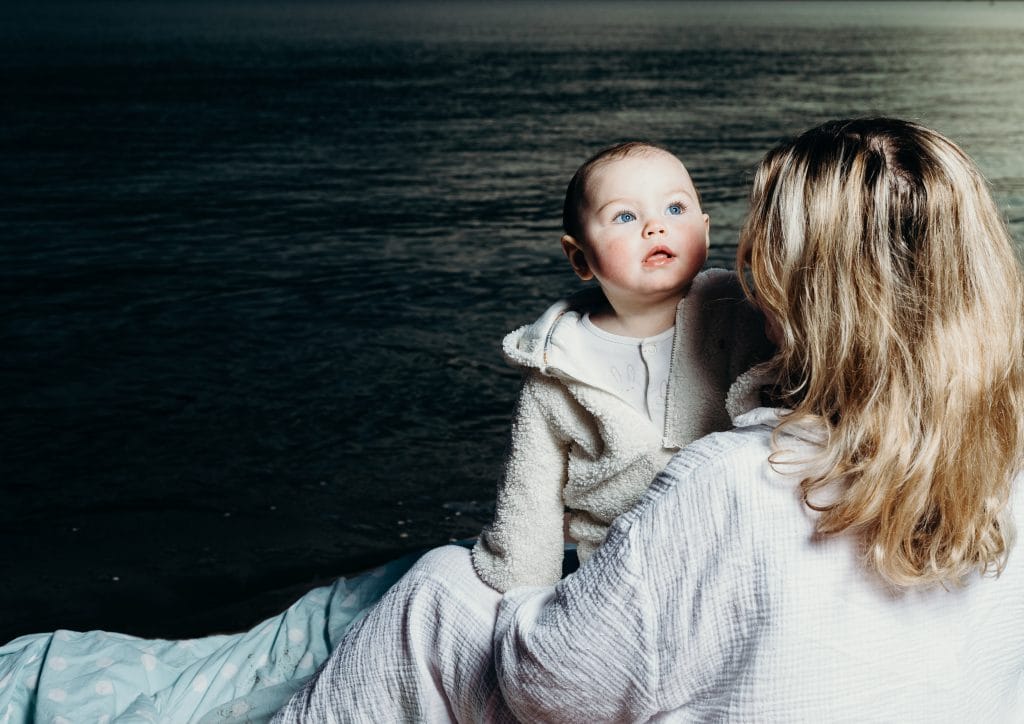 A photo of a baby looking over their parents shoulder. In the background there is a dark sea